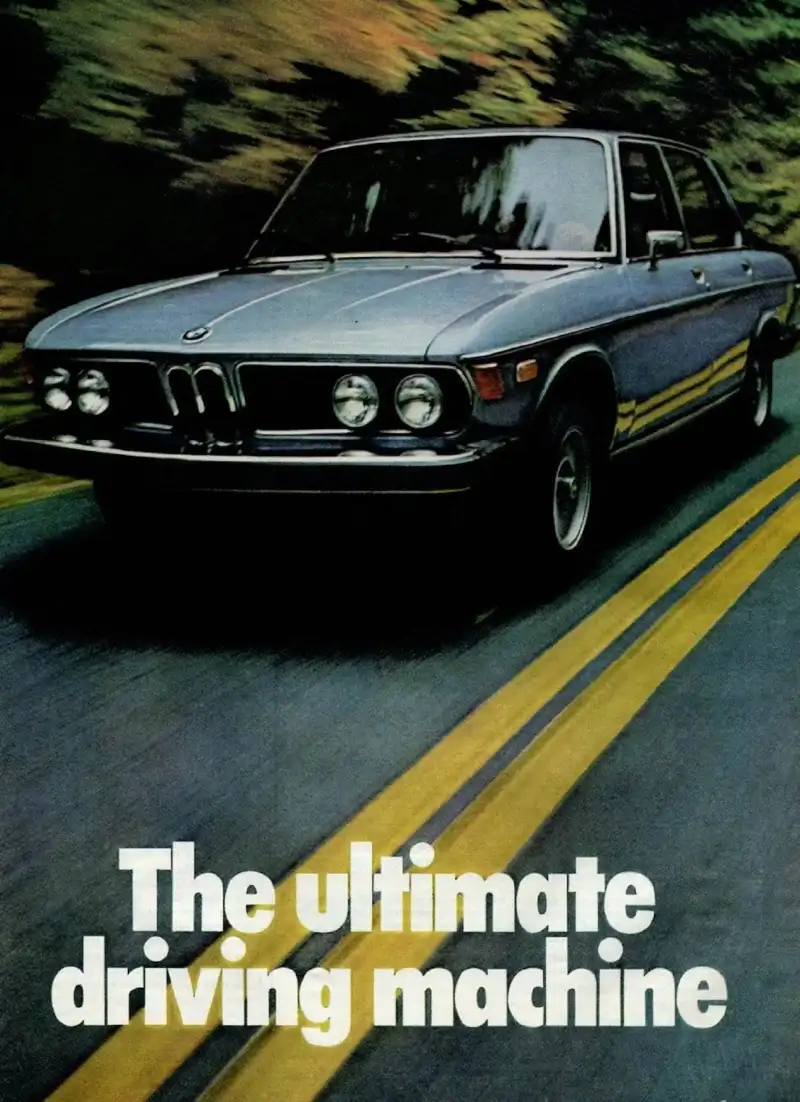 A vintage light blue BMW sedan, seen from the front three-quarter angle, driving on a dark asphalt road with two bright yellow lines. The background is a blurry wash of green and brown foliage, suggesting motion. White text at the bottom reads 'The ultimate driving machine'.